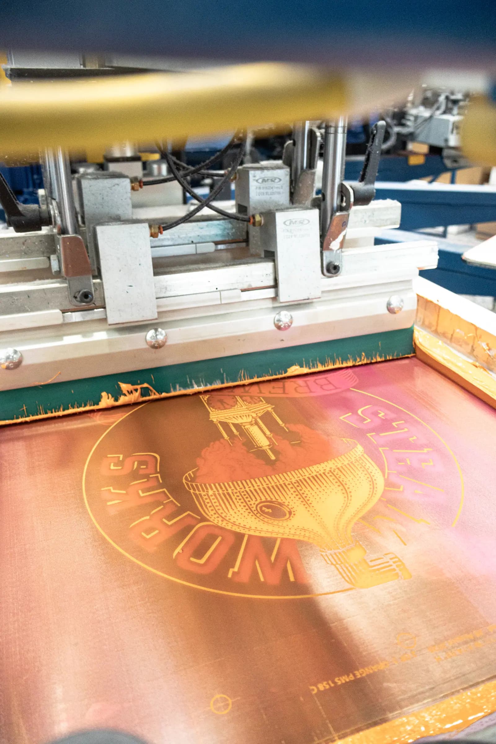 Orange ink pulled across a screen on the press, with a Pantone reference on the screen edge